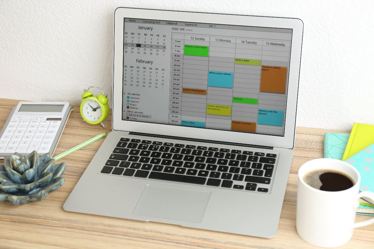 Laptop displaying a digital calendar schedule on a desk with office supplies and a cup of coffee