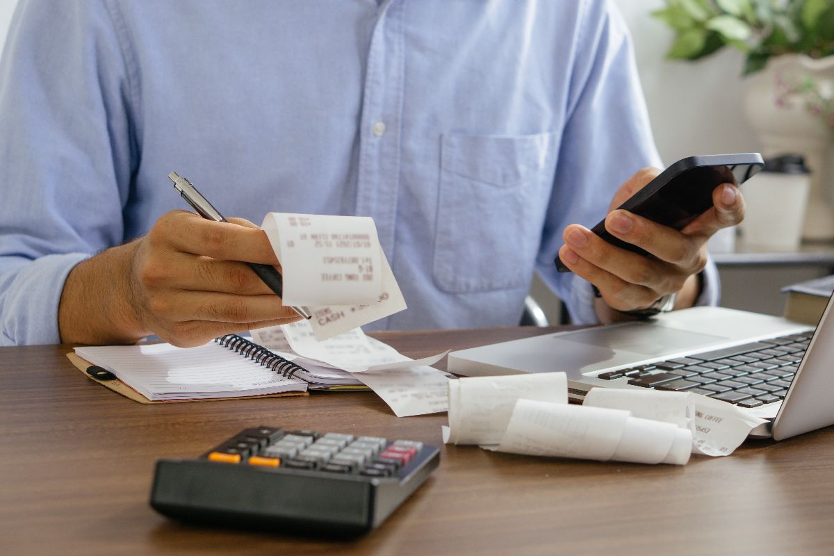 Person reviewing receipts and using a smartphone beside a calculator and laptop for manual accounting tasks
